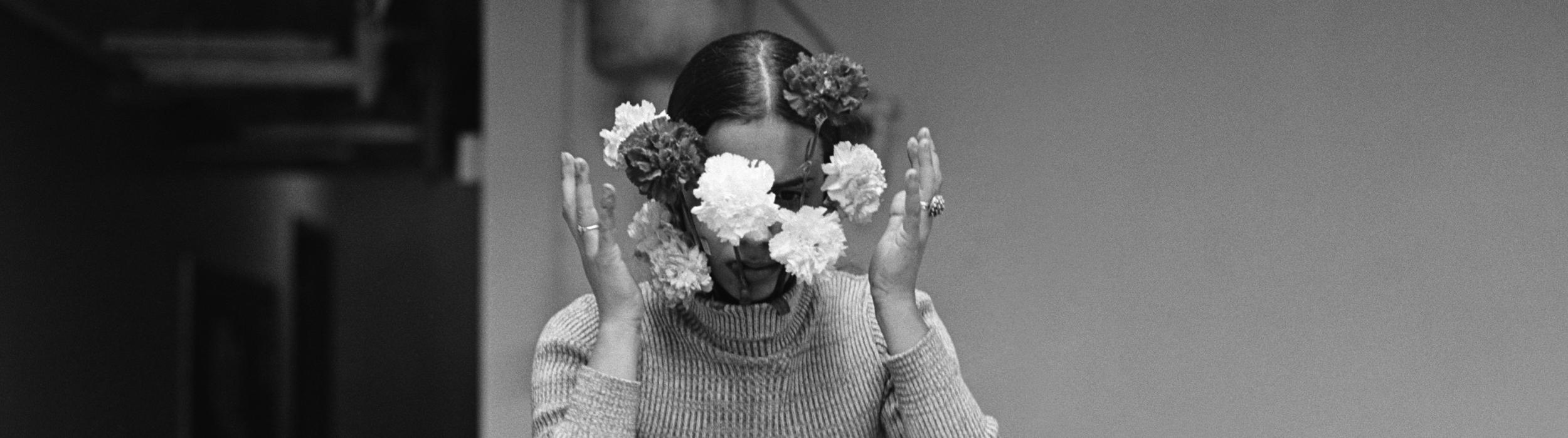 Photo noir et blanc d'une femme, mains légèrement écartées du visage, tournée vers nous et dont on perçoit à peine le regard, avec des fleurs blanches et d'autres plus sombres tout autour de la tête.